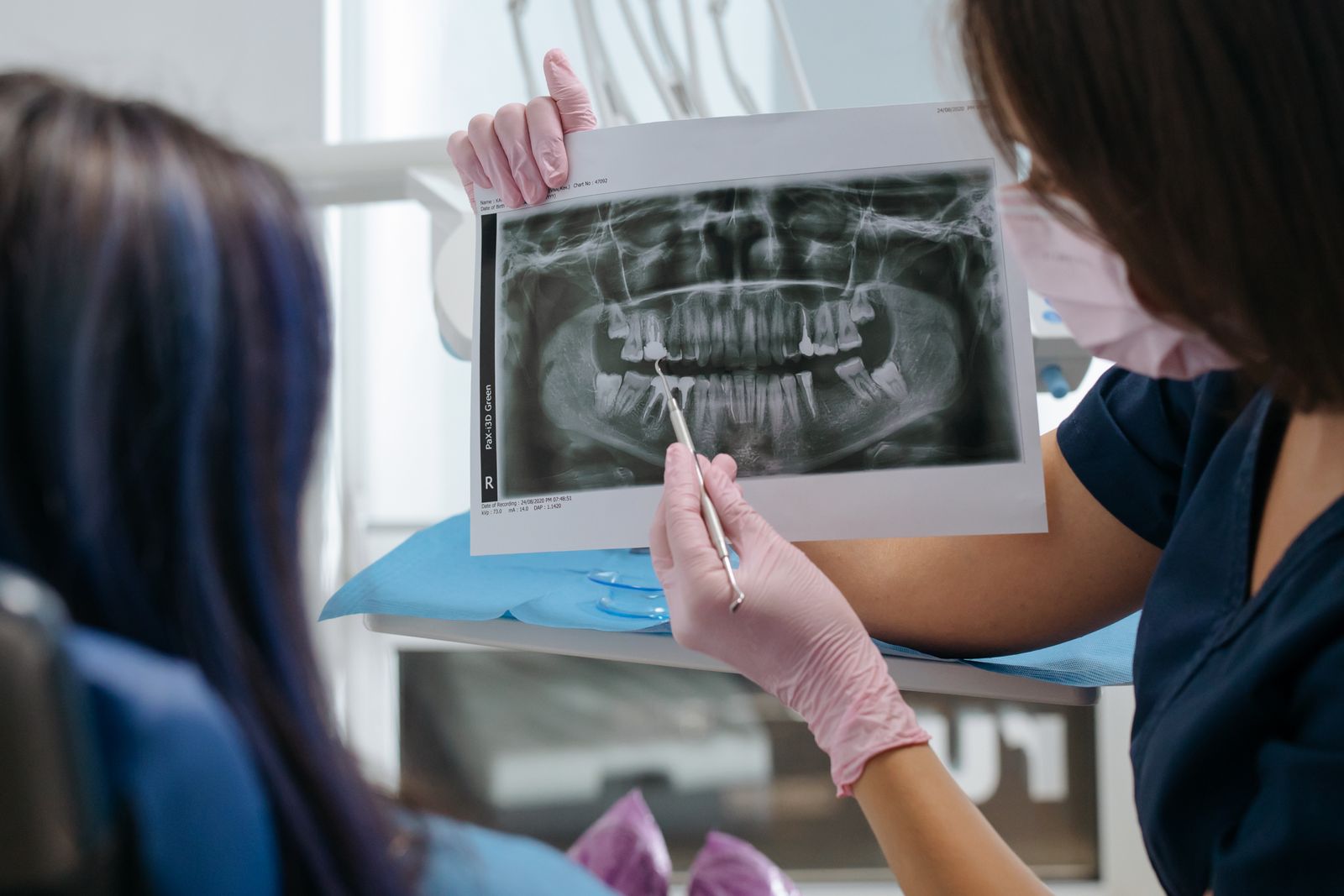 Dentista con mascarilla y guantes sostiene una radiografía panorámica y señala con un instrumento la raíz de un diente mientras lo explica a una paciente en el sillón